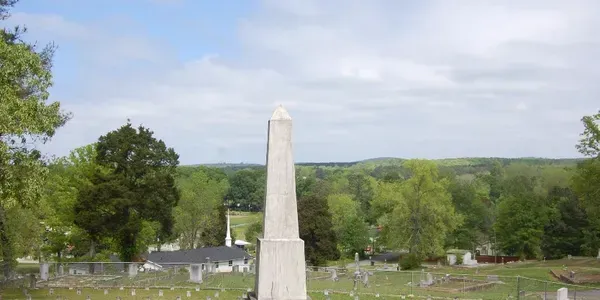 Tall white obelisk monument in a green park with trees and hills under a cloudy sky
