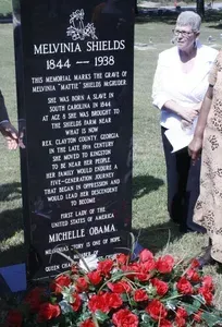 Memorial plaque for Melvinia Shields beside a person holding papers, with red flowers at the base