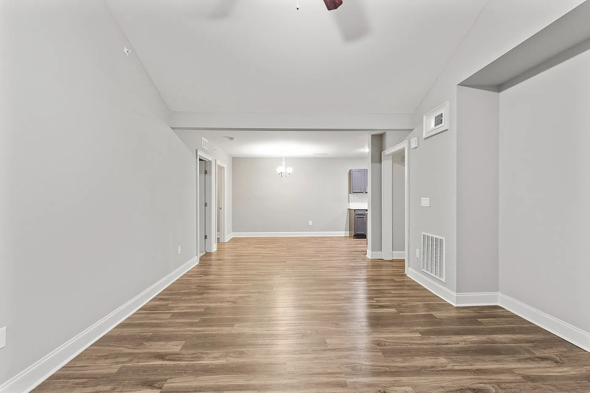 Empty living area in a modern apartment with wood-like flooring and a view of the kitchen.