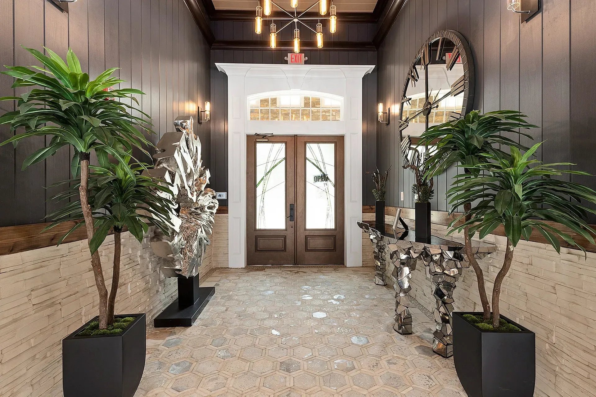 Lobby entrance with double wooden doors, pendant chandelier, and large potted plants flanking the doorway.