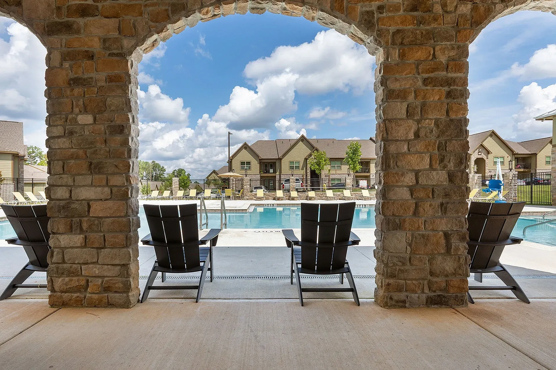 Stone archway frames a resort-style pool with lounge chairs and apartment buildings in the background.