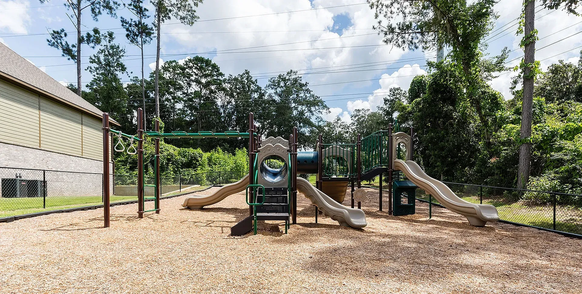 Outdoor playground with slides and climbing structures on wood-chip ground, fenced, with trees in background.
