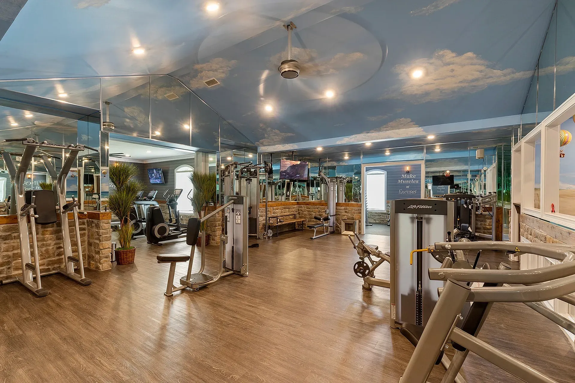Light-filled fitness center with cardio and strength machines, mirrors, and wood-look flooring.