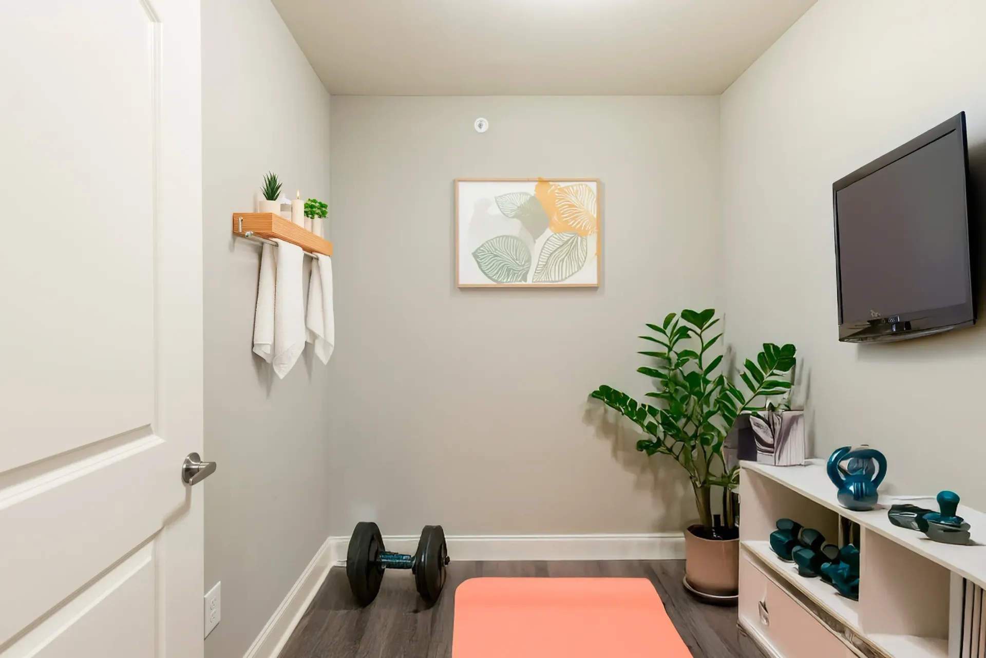 Small fitness room with a yoga mat, dumbbells, towels on a shelf, and a wall-mounted TV.