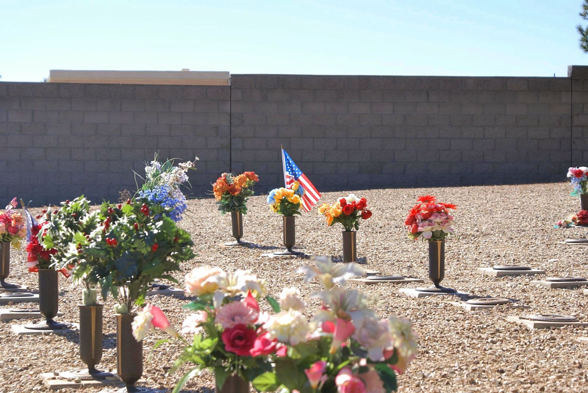 Graveyard with colorful flowers in vases and a U.S. flag, set against a gray wall under a clear sky.