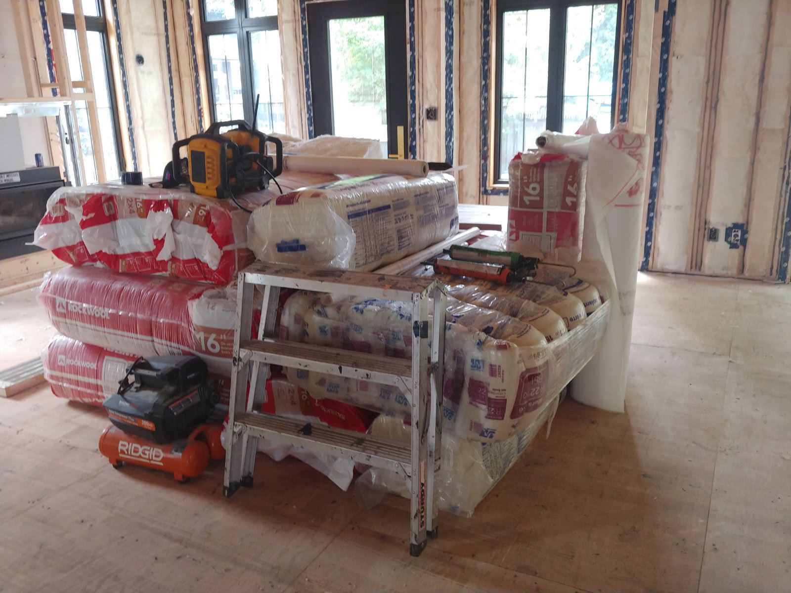 Stacked construction materials on a pallet in an unfinished room with exposed walls and windows.