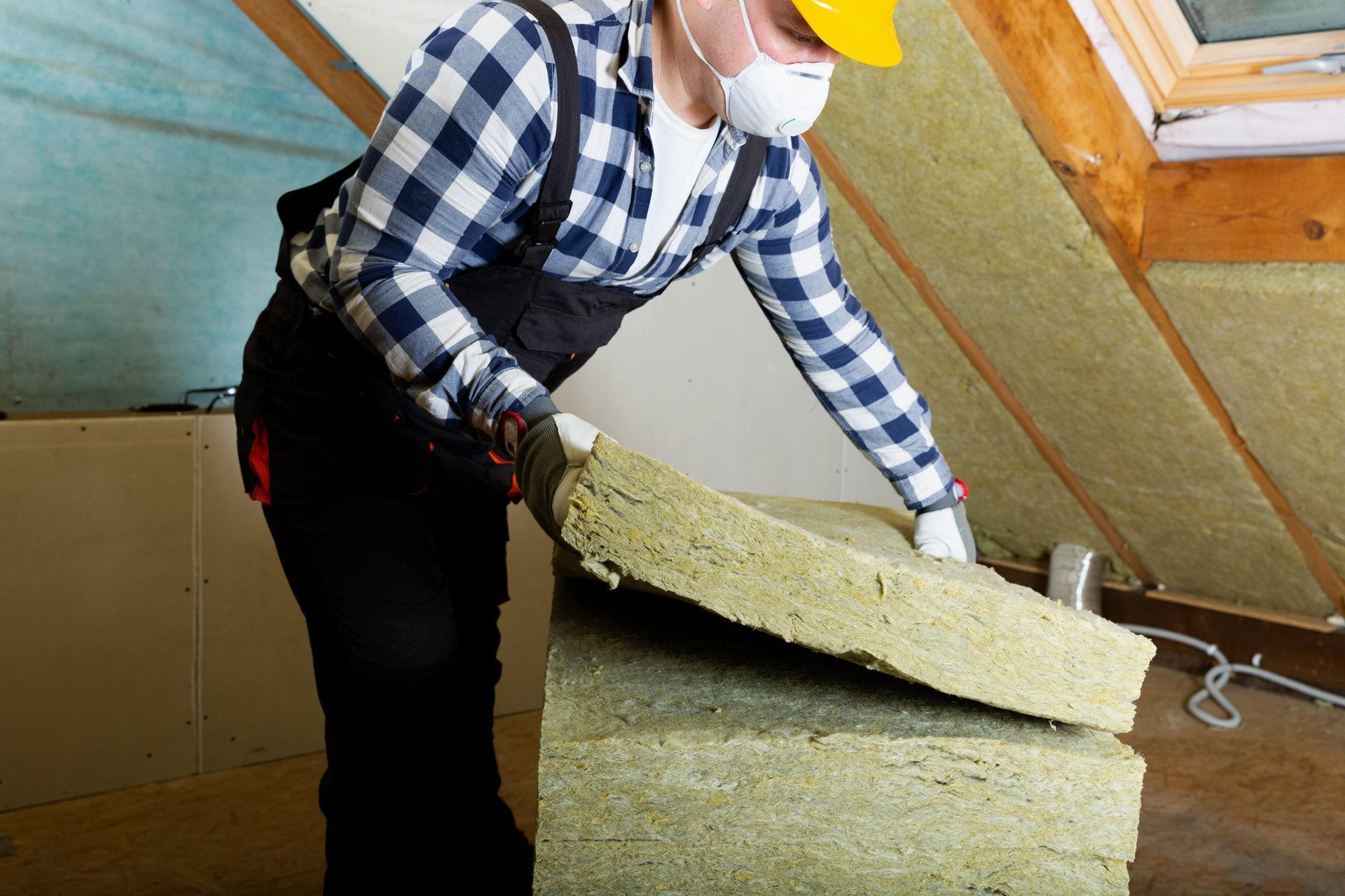 Worker in a yellow hard hat lifting a large insulation panel in an attic.