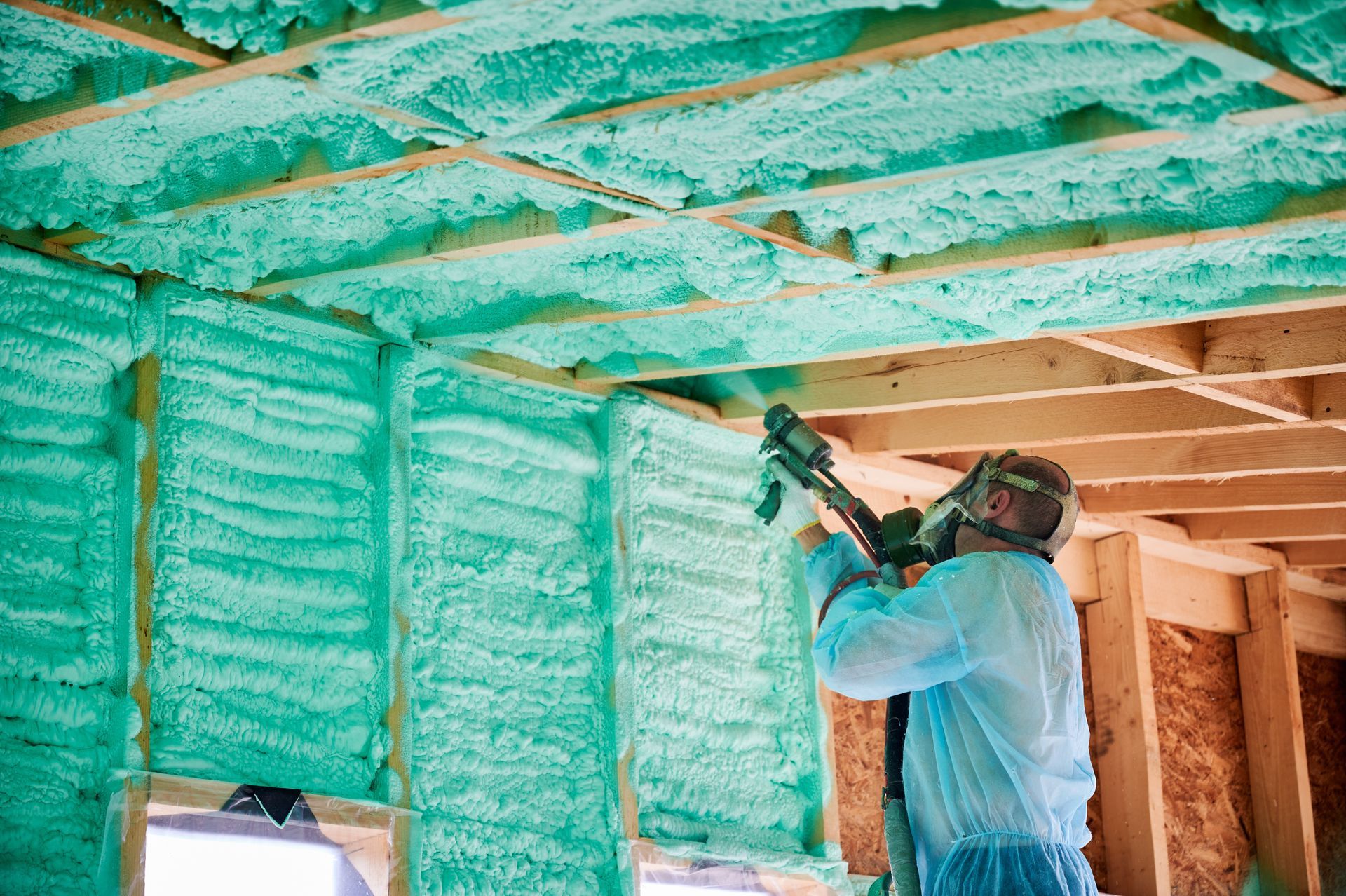 Worker spraying green foam insulation on walls and ceiling in a house under construction