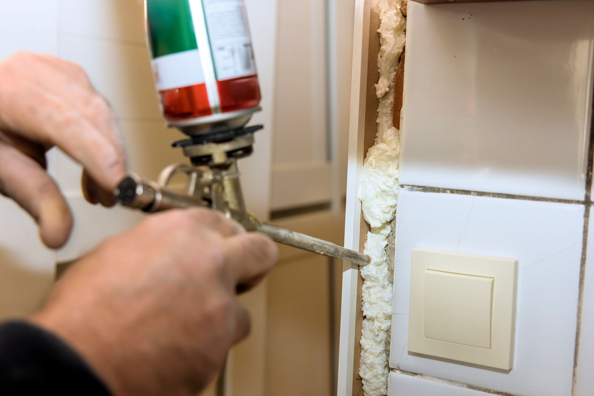 Hands using a spray foam gun to insulate a gap beside a wall and light switch.