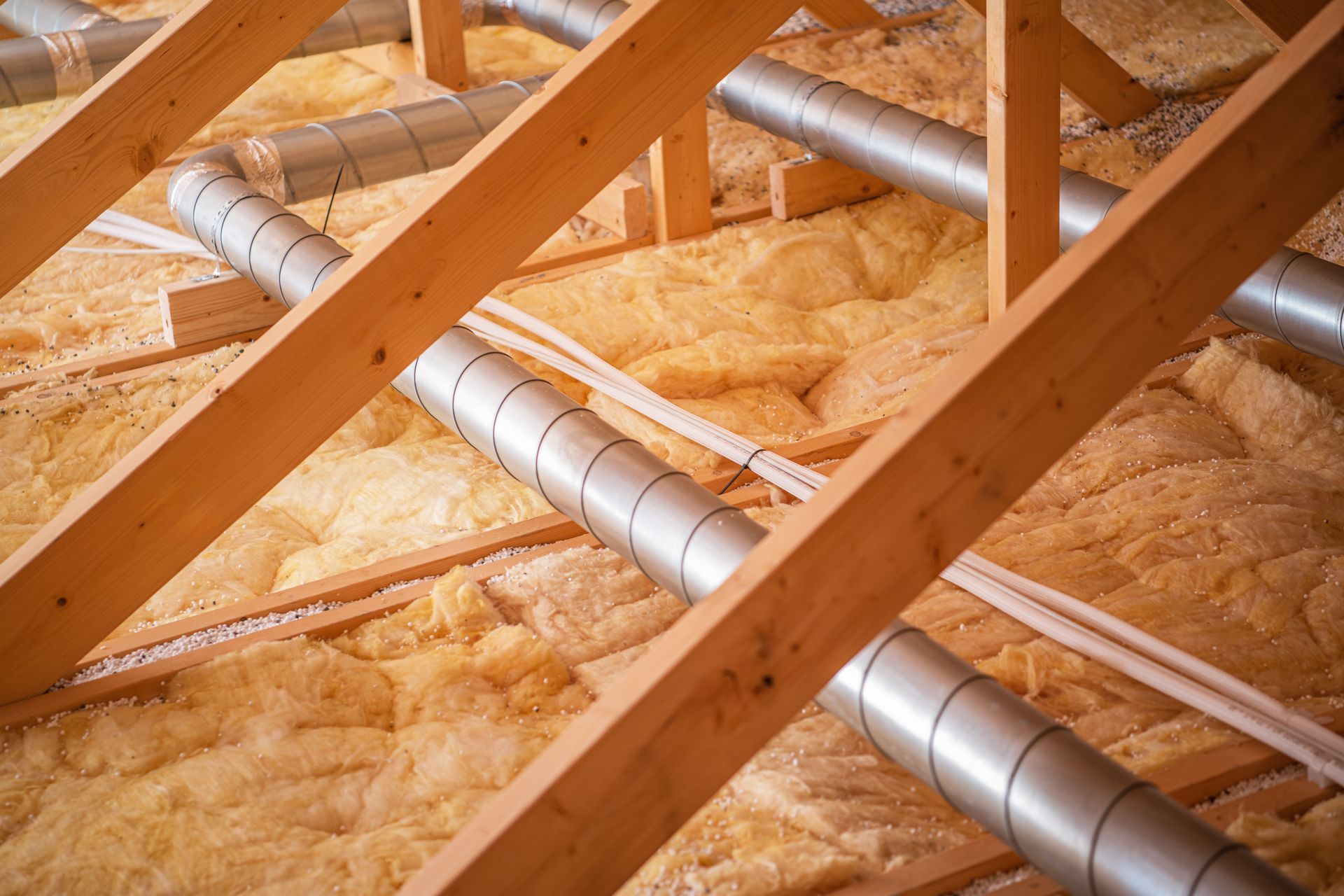Wooden roof trusses over orange insulation with silver ducting in an unfinished attic