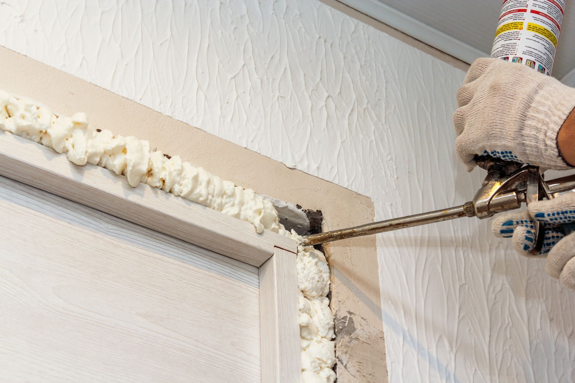Foam insulation being sprayed into a gap around a window frame with a caulking gun attached to a canister