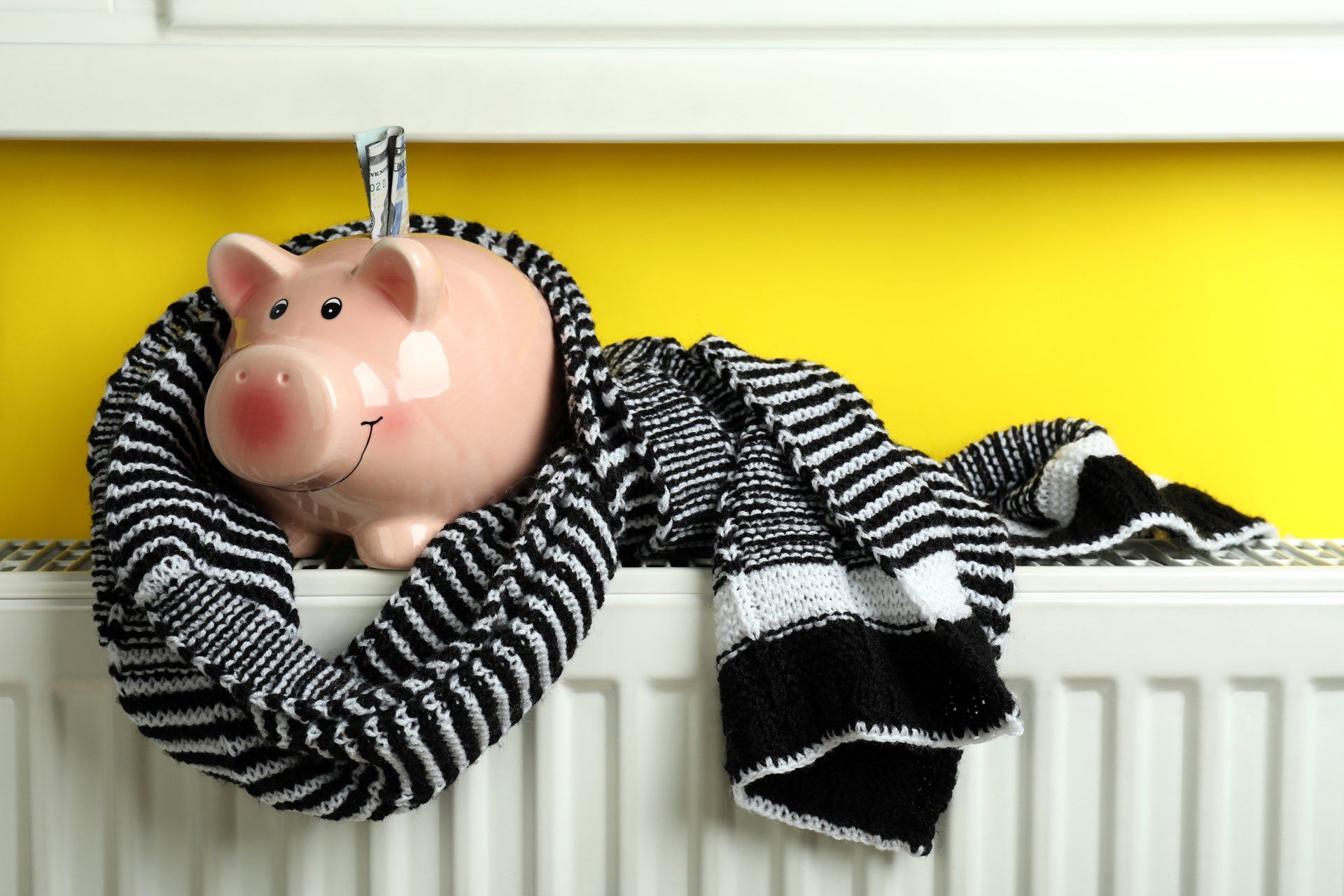 Pink piggy bank draped in a black-and-white striped scarf on a white radiator with a yellow wall behind