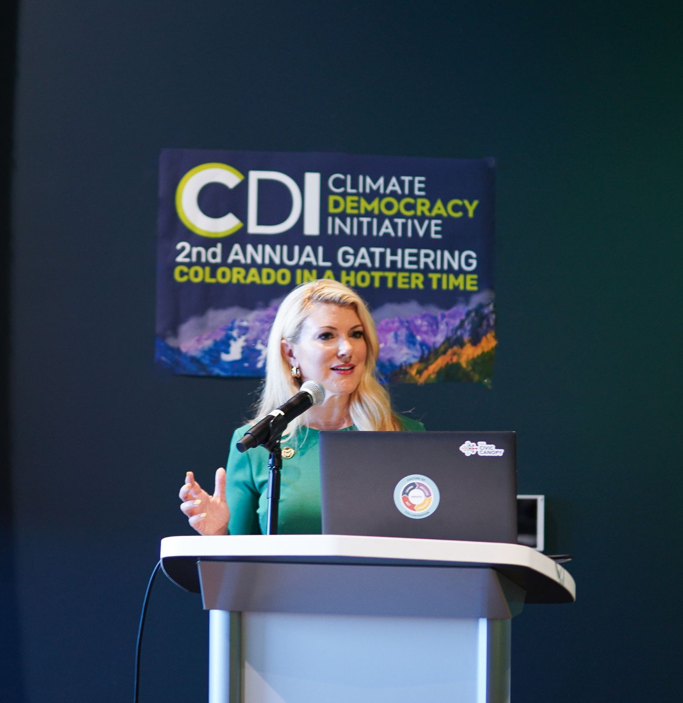 Speaker at podium on climate democracy event stage, with “CDI” banner in the background.