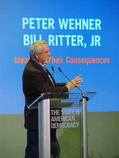 Man speaking at a podium onstage before a blue screen reading “Peter Wehner” and “Bill Ritter, Jr.”