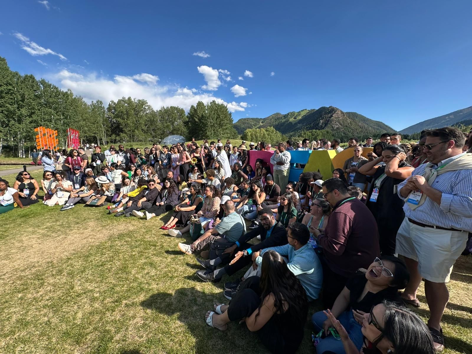 Crowd seated on grass at an outdoor event under a blue sky, with trees and hills in the background