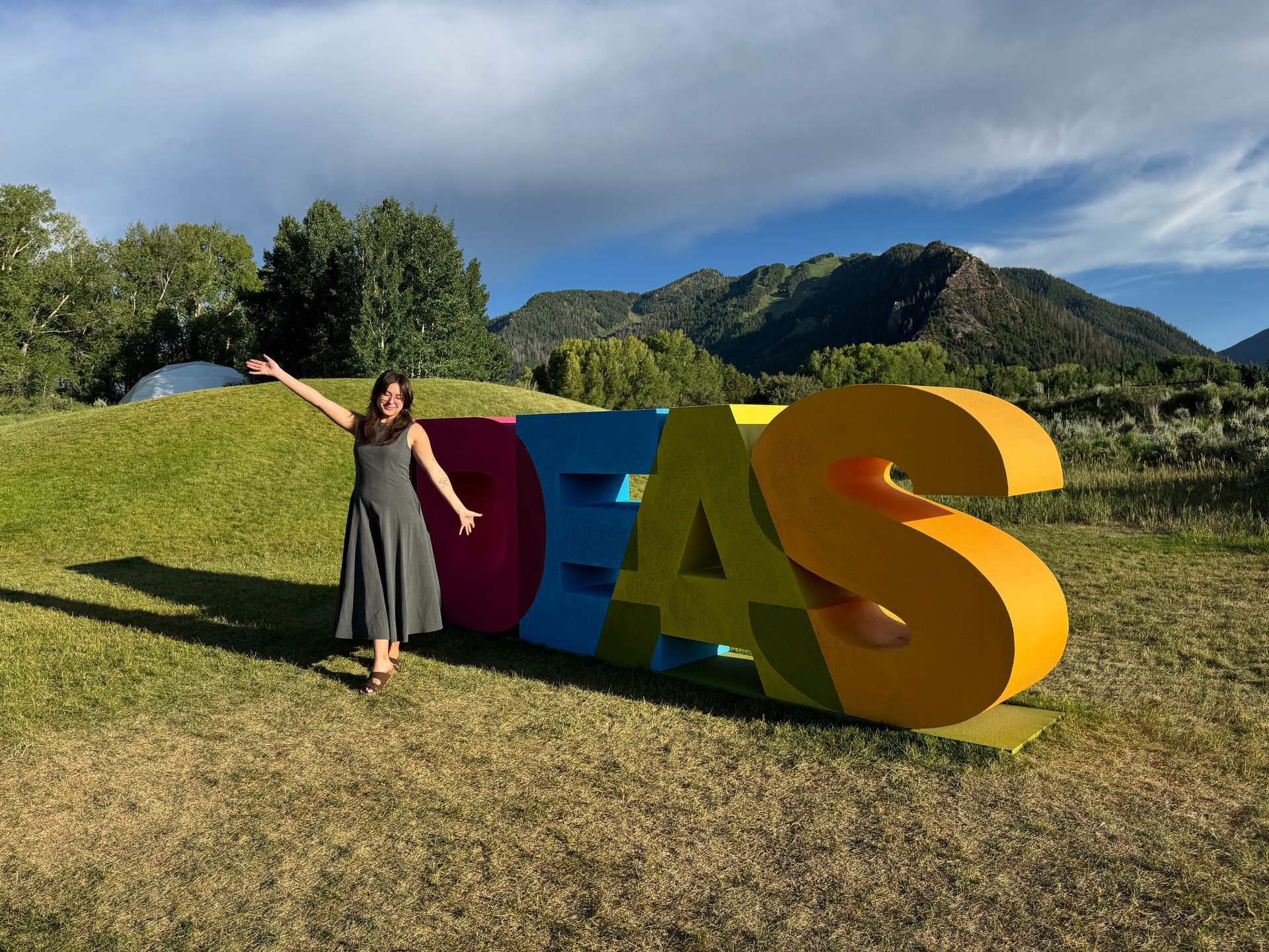 Woman posing beside large colorful letters on a grassy hill with mountains and blue sky in the background