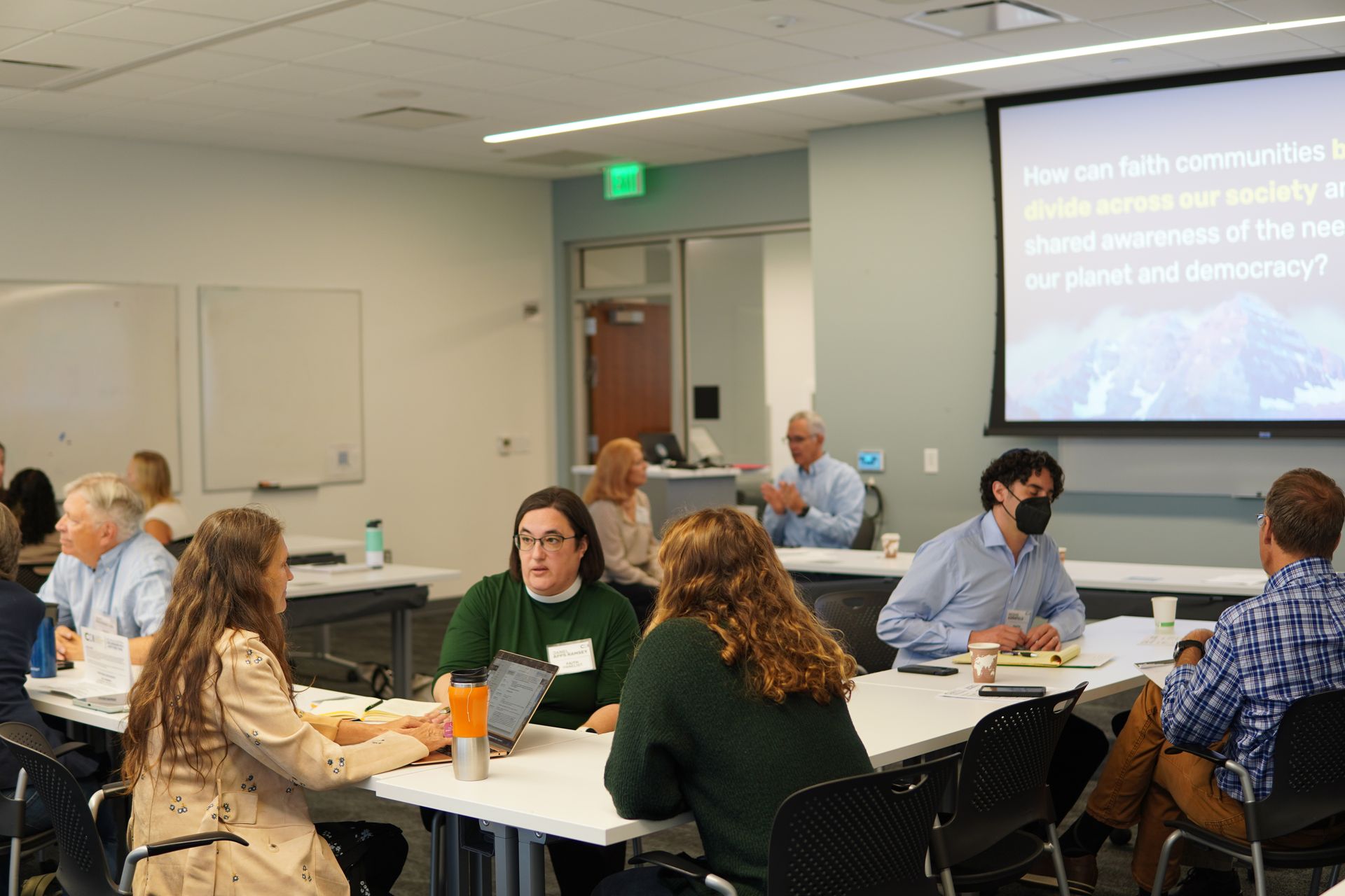 People seated around tables in a classroom during a group discussion with a projected presentation.