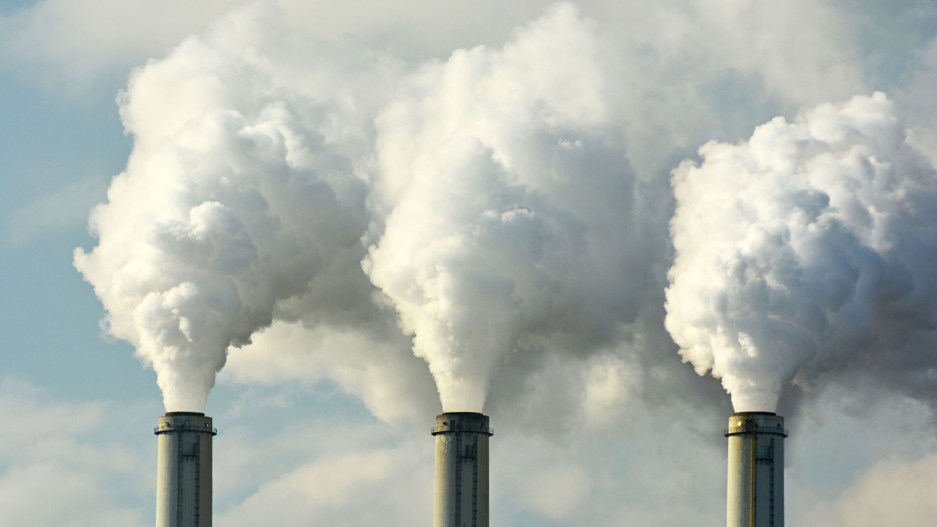Three smokestacks release thick white smoke into a cloudy sky.