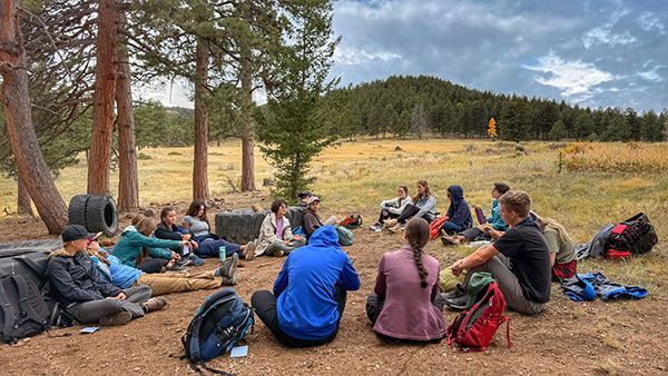 Group of people sitting on logs in a forest clearing, facing a speaker during an outdoor gathering