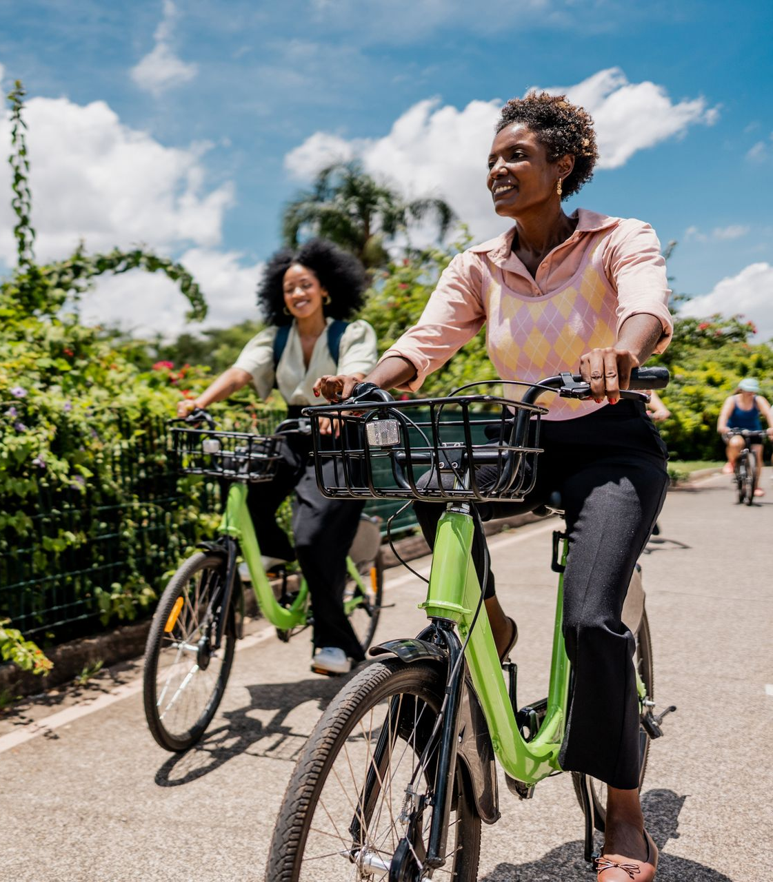 Two people ride bicycles on a sunny path, with green hedges and blue sky in the background.