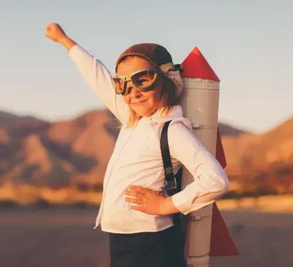 Person in goggles and a white shirt posing with a rocket backpack in a desert landscape, arm raised.