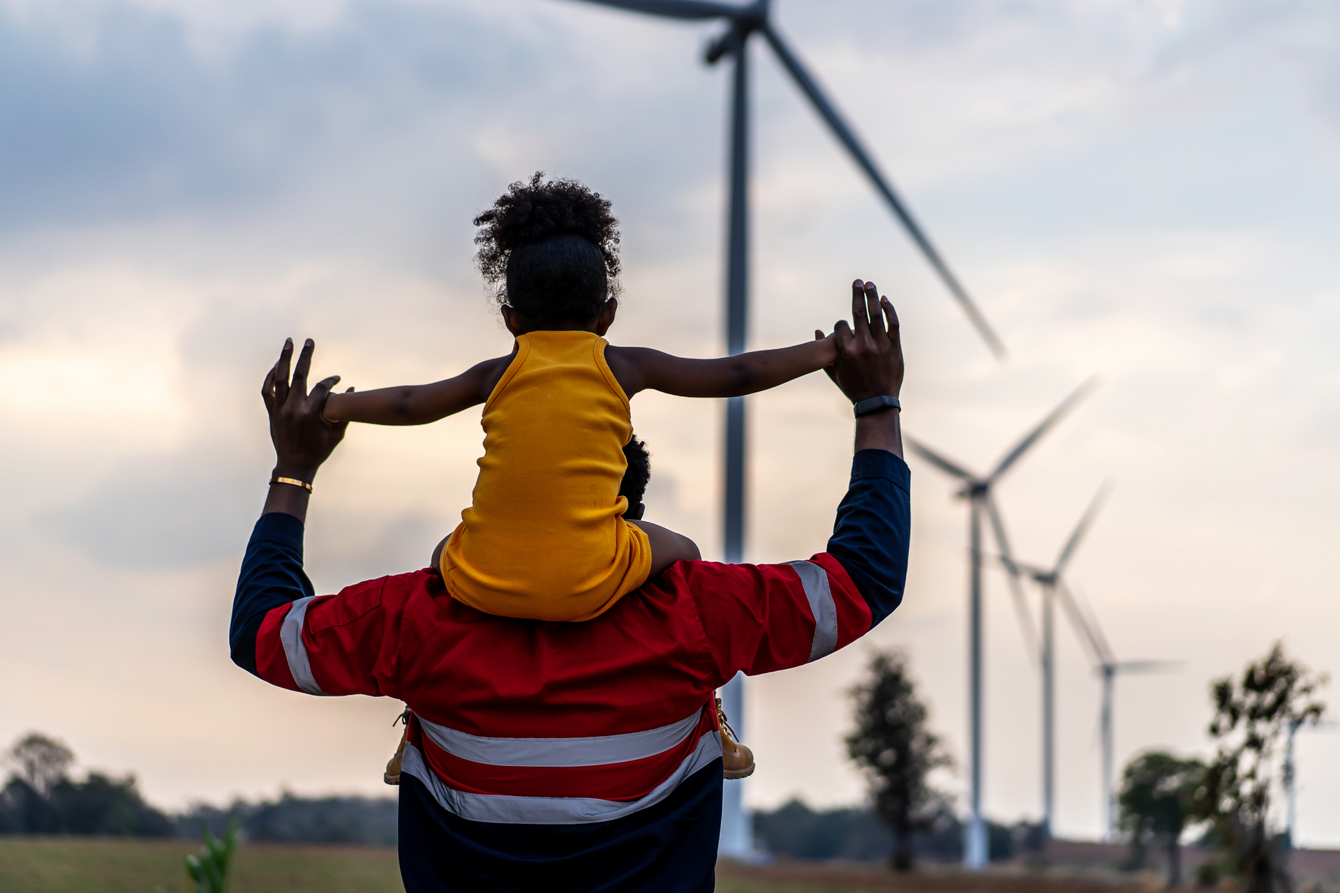 Adult carrying child on shoulders in a field with wind turbines at sunset