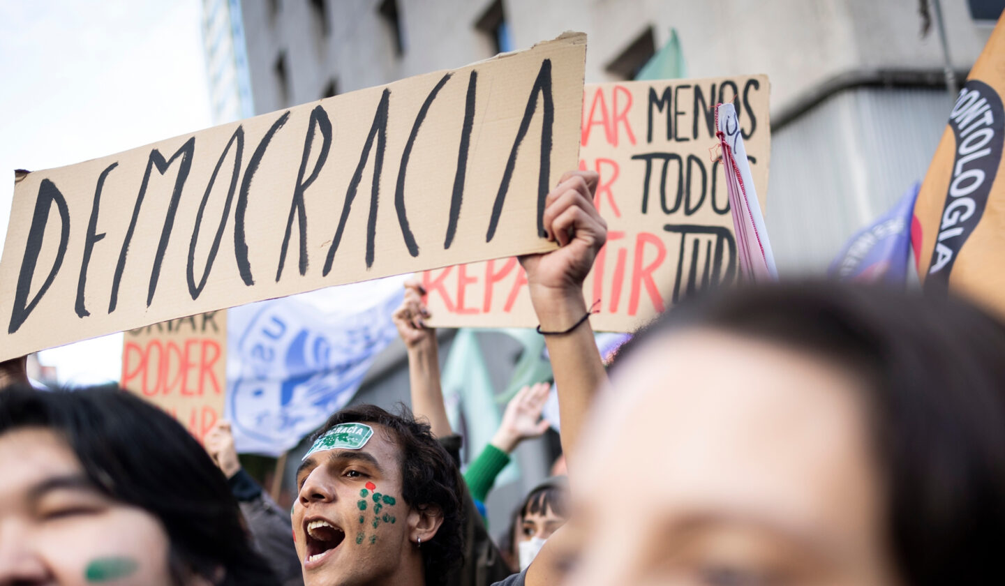 Protesters holding signs, including “DEMOCRACIA,” in a crowded street demonstration