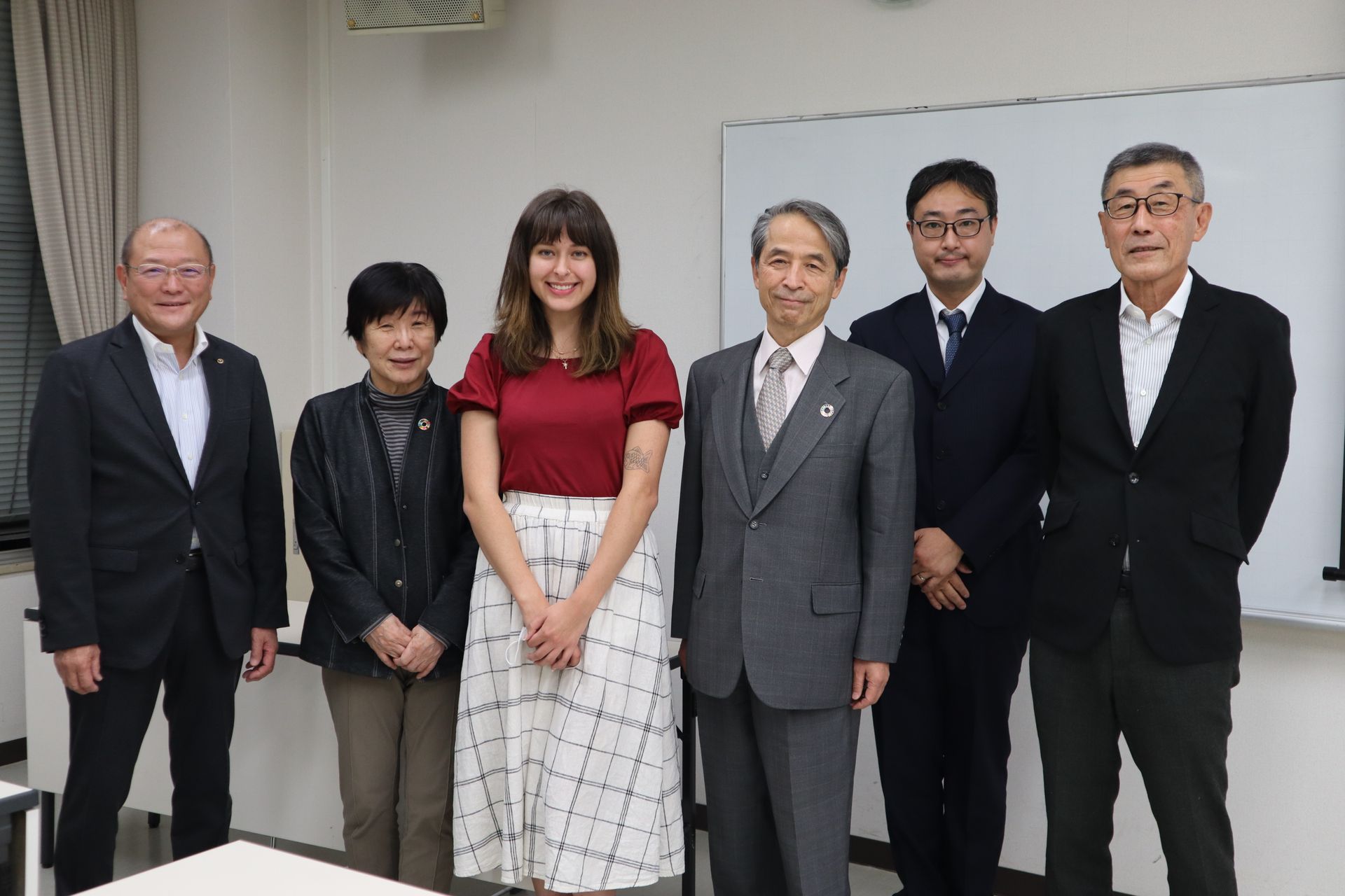 Five people standing indoors against a plain wall, dressed in formal attire, posing for a group photo