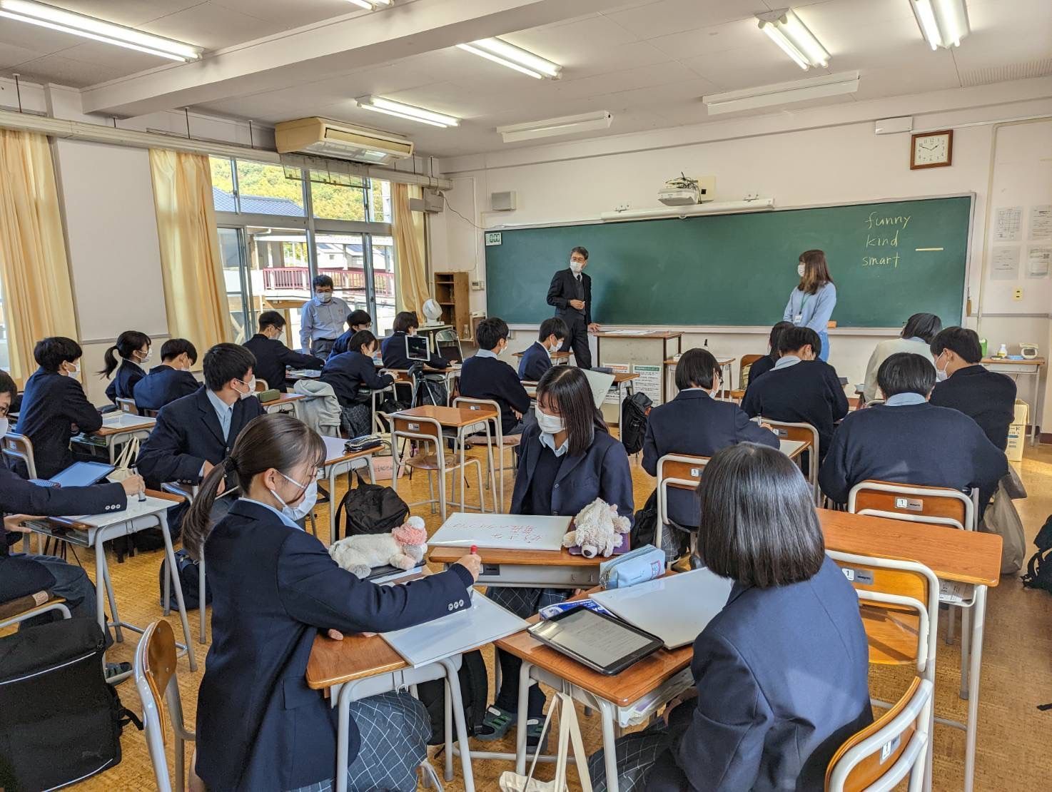 Classroom with students seated at desks, teachers at the chalkboard, and large windows on the left.