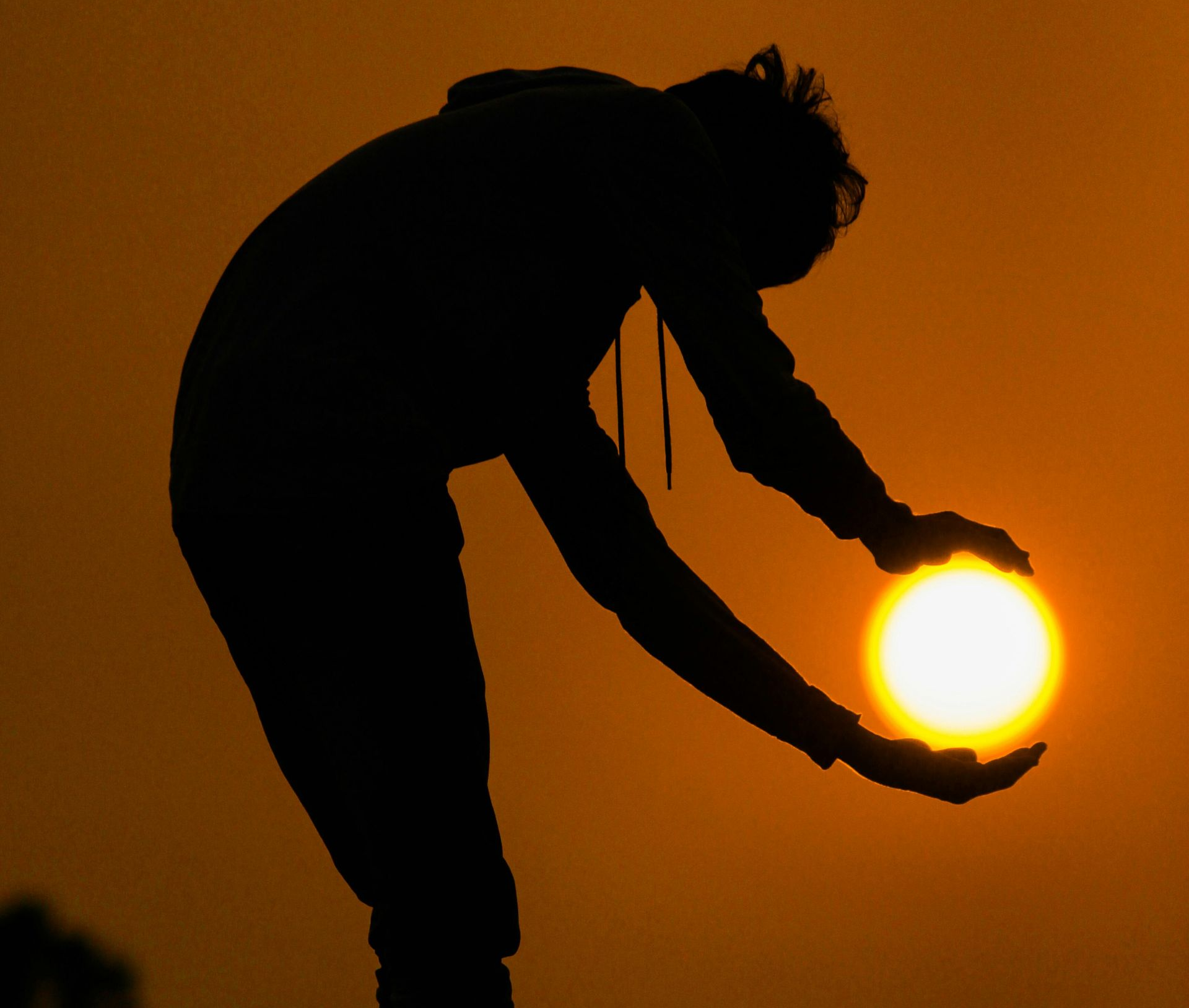 Silhouette of a person holding the sun at sunset against an orange sky