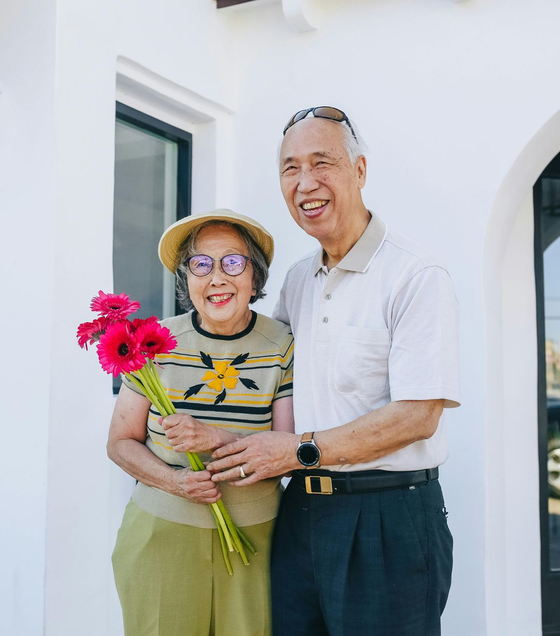 Smiling couple standing outdoors, holding a bouquet of bright pink flowers near a white house