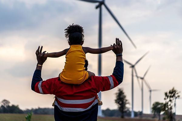 Person carrying a child on shoulders near wind turbines at sunset