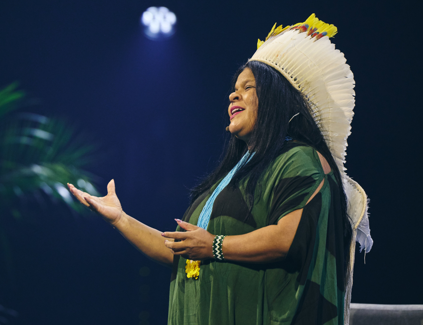 Singer in a green dress and feathered headdress performing on stage with arms outstretched under spotlights