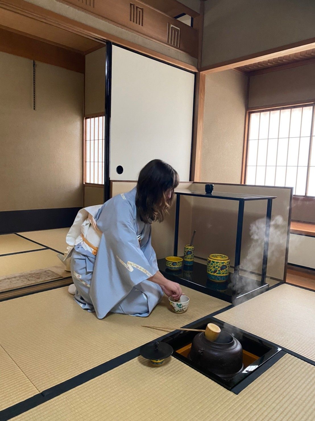 Person kneels in a tatami room, arranging flowers beside a black kettle and tea setup.
