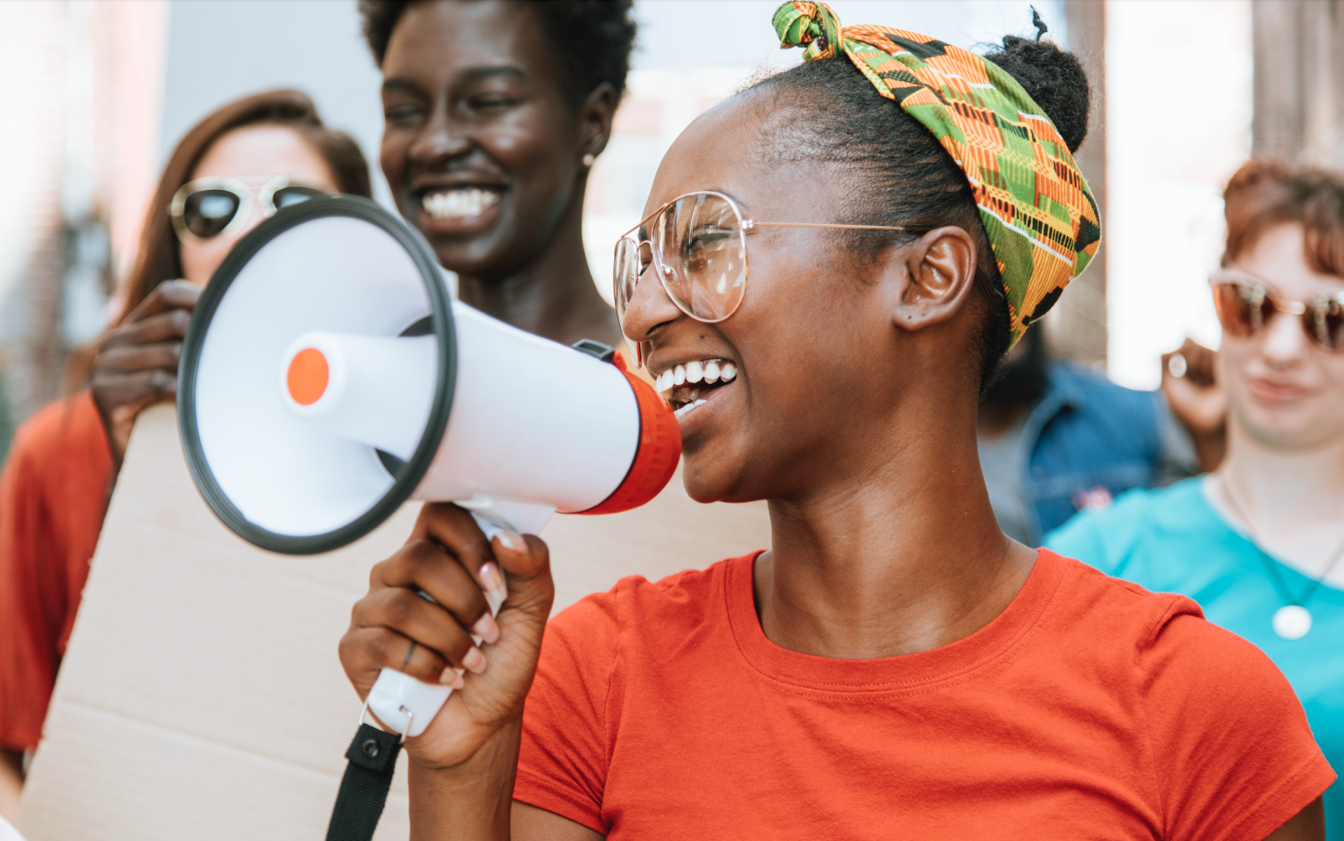 Person speaking into a megaphone at a rally, wearing an orange shirt and colorful headscarf.