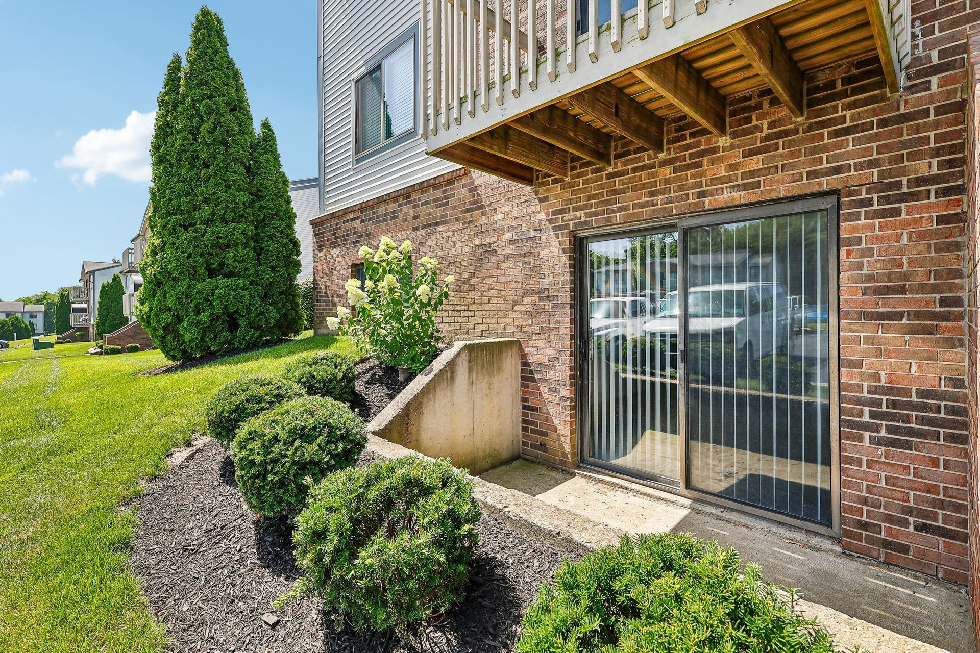 Tall Timber Apartments Brick apartment exterior with sliding glass door, stairs, and landscaped shrubs along a walkway