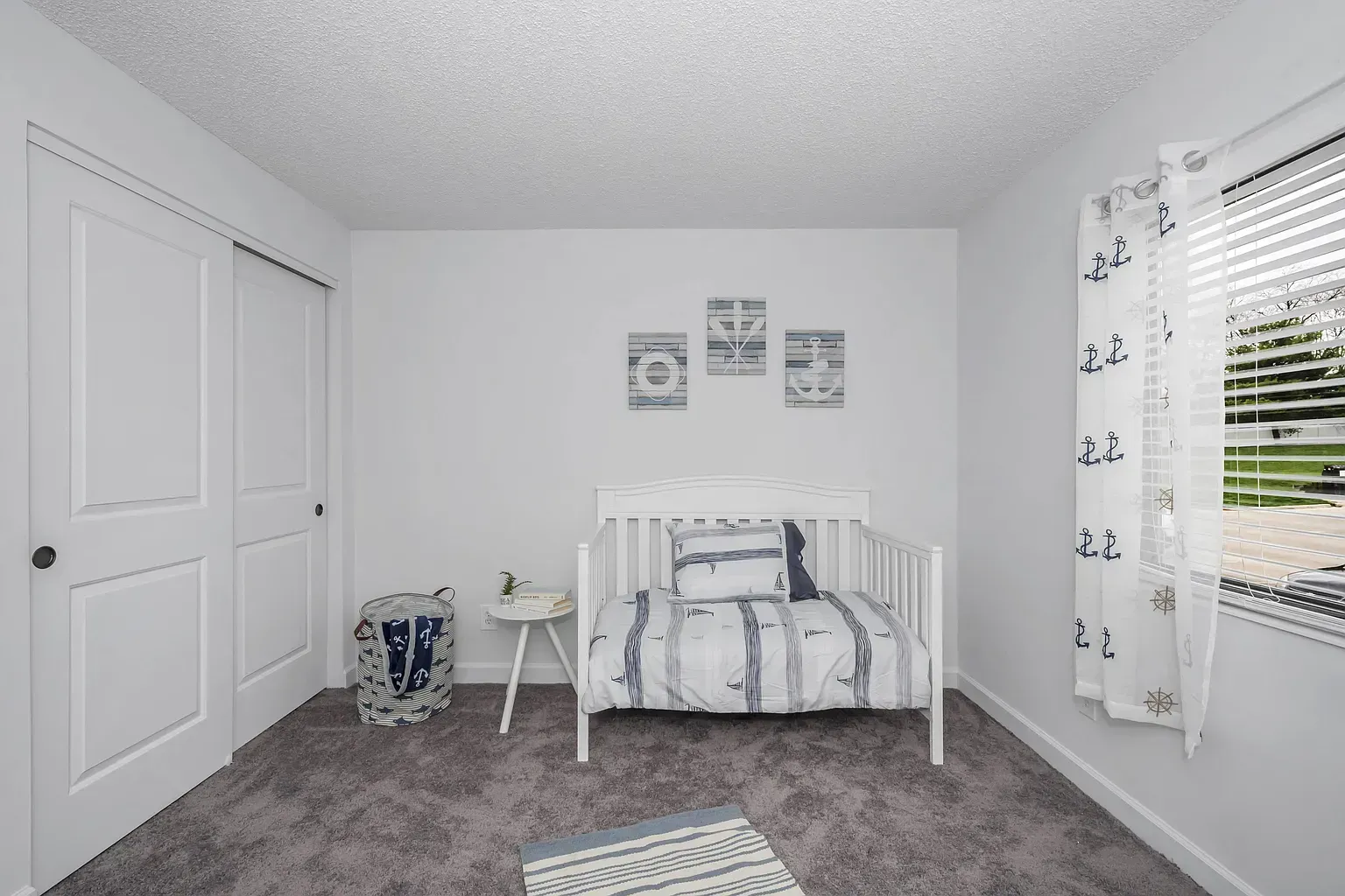 Tall Timber Apartments Small white toddler bed in a bright bedroom with a window, striped bedding, and nautical decor.
