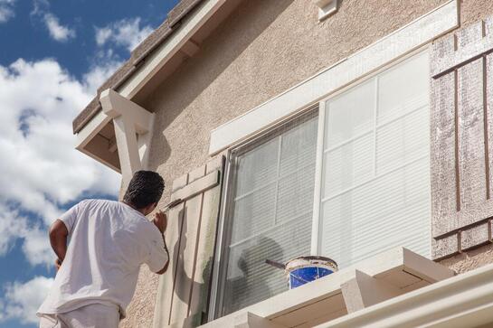 Person painting the exterior of a beige house under a blue sky
