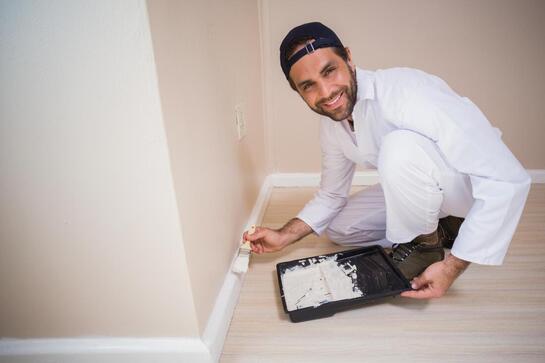 Painter kneeling by a wall, applying white paint with a brush and tray on a wooden floor