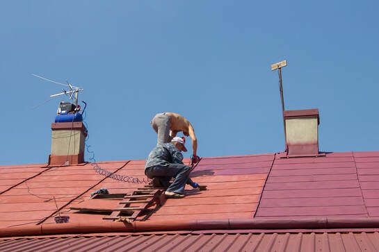 Two workers on a red roof installing shingles under a clear blue sky