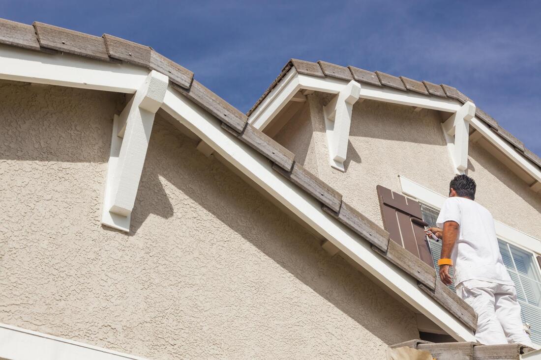Worker painting a house exterior while standing on a ladder near the roofline