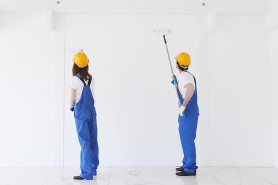 Two workers in blue coveralls and yellow hard hats paint a white wall with rollers.
