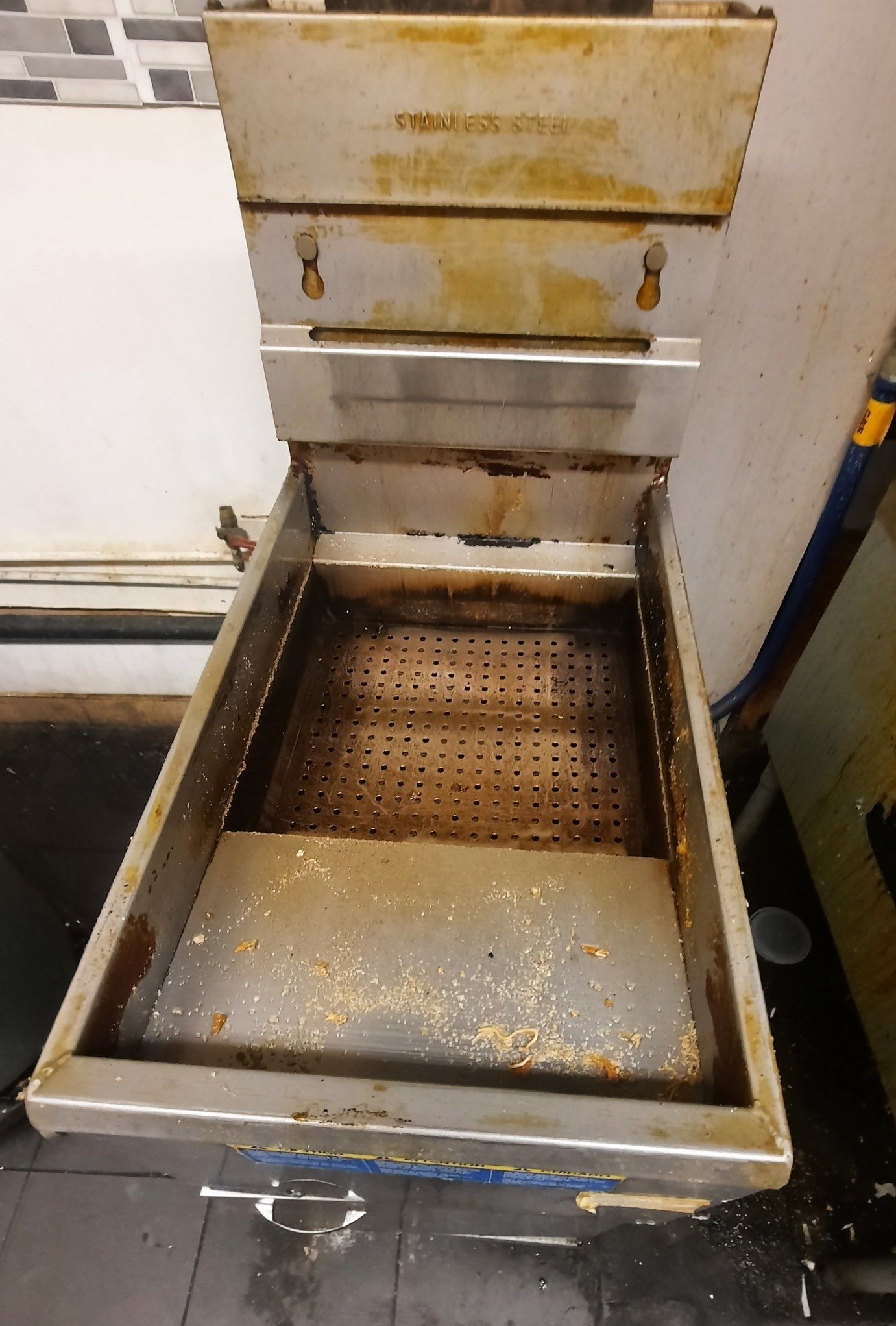 Une friteuse en acier inoxydable sale repose sur un sol carrelé.