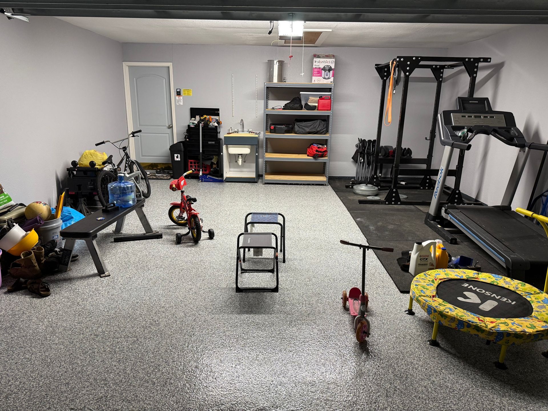 Garage gym with fitness equipment, kids' toys, and storage shelves. Gray walls and flooring.