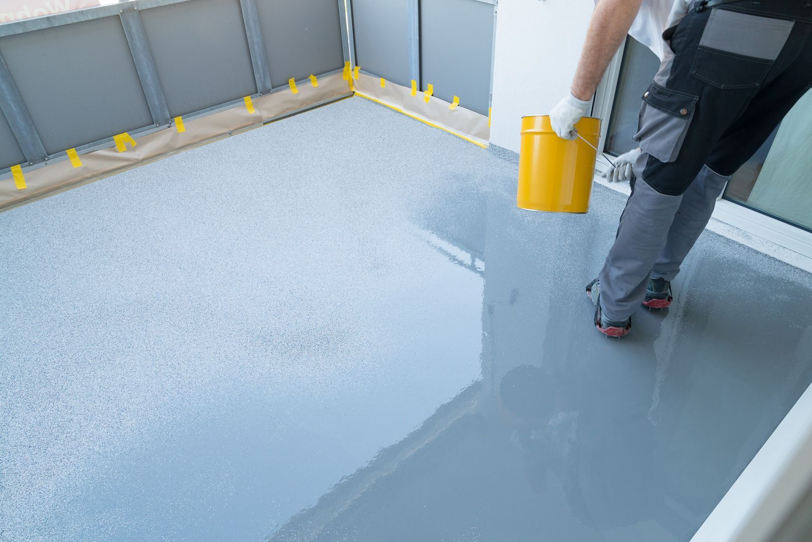 Person pouring gray epoxy coating on a floor, with a yellow bucket.