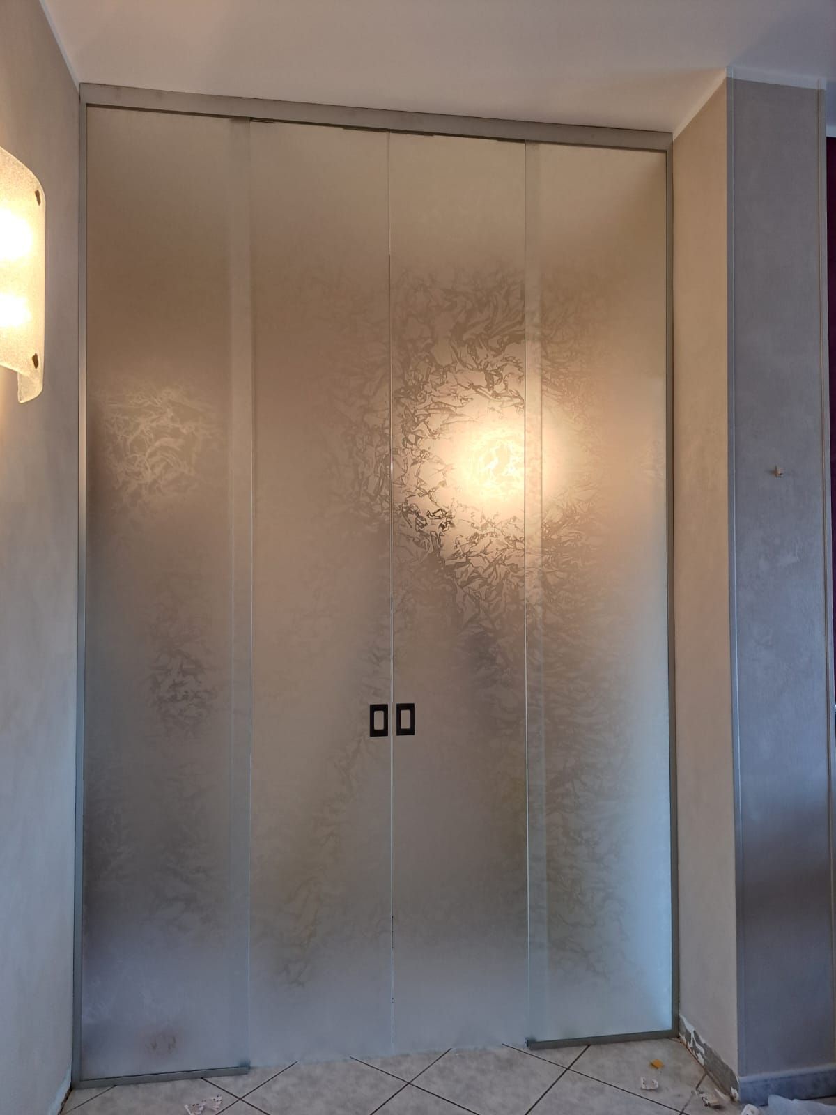 Doppie porte in vetro satinato in un corridoio con applique a muro e pavimento piastrellato.