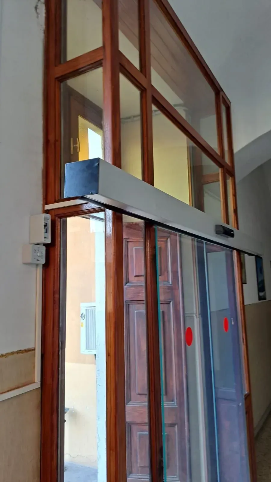 Un ascensore stretto con pareti in vetro incorniciate in legno e porte scorrevoli in metallo in un corridoio.