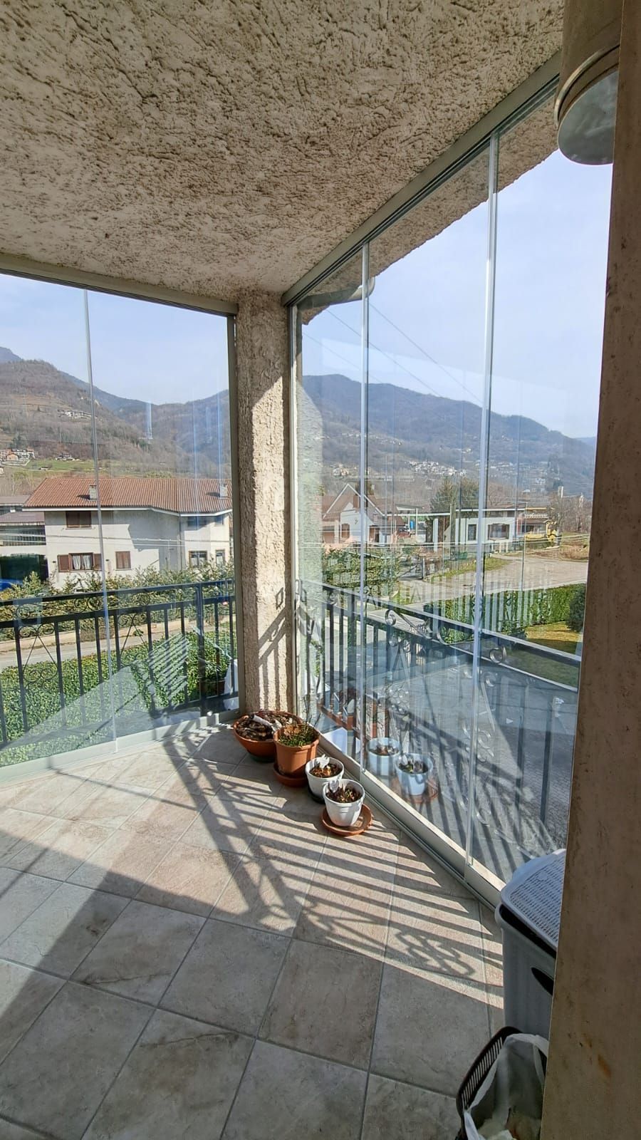 Veranda coperta con pavimento piastrellato, piante in vaso e vista sulle montagne attraverso la ringhiera e i pannelli di vetro.