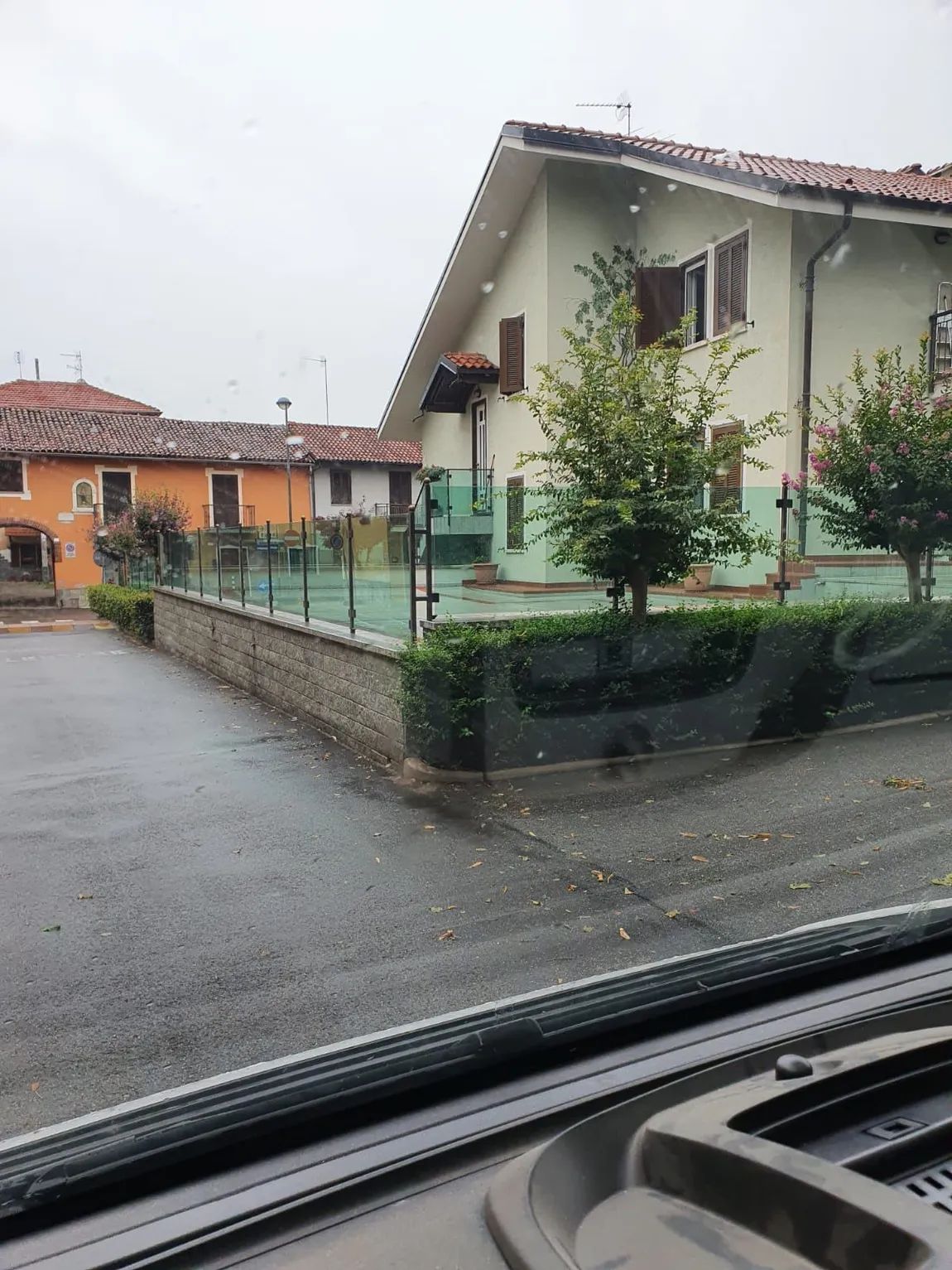 Strada residenziale piovosa vista da un'auto, con case, un muretto basso e un furgone arancione parcheggiato.