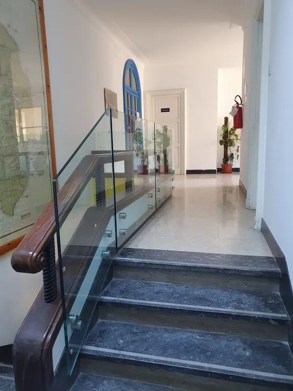 Scala interna con ringhiera in vetro che conduce a un corridoio luminoso con piante in vaso.